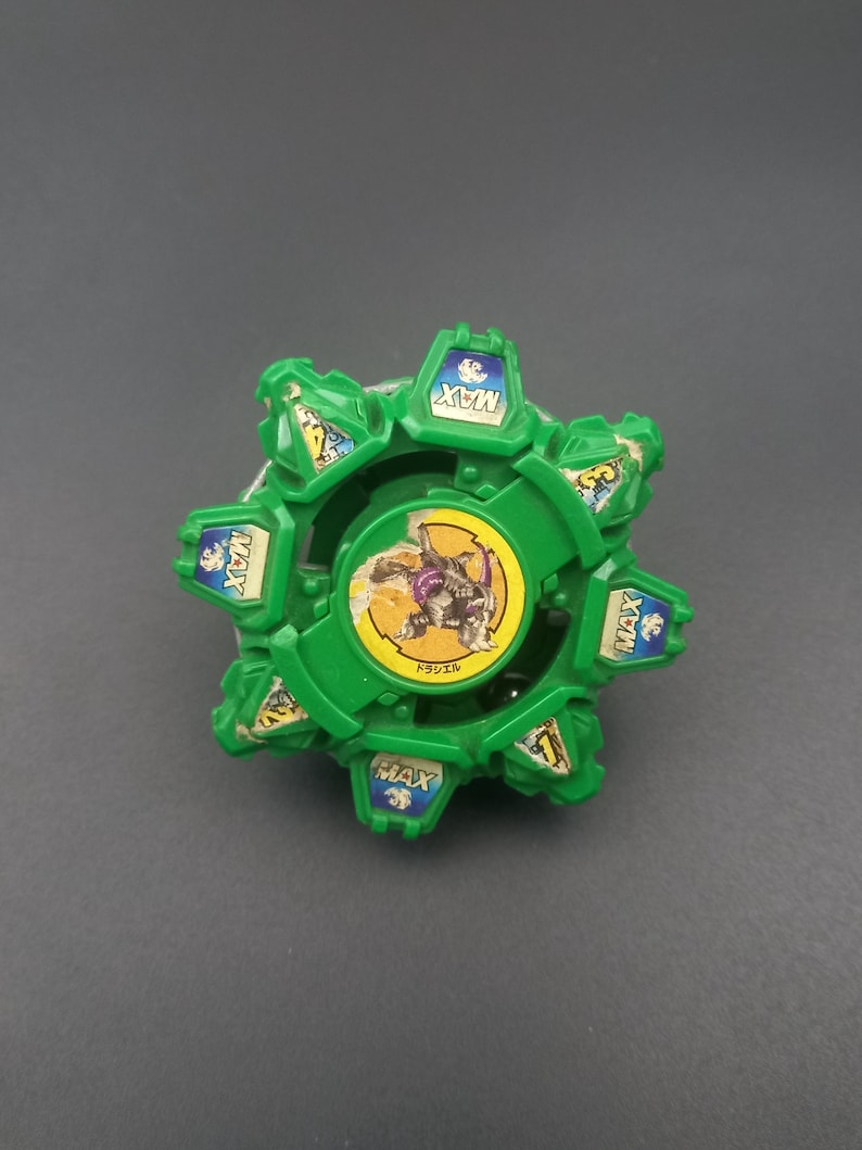 May include: A green and blue toy with a circular design. The toy has a central image of a character and the word "MAX" on several sections. The toy is on a gray surface.