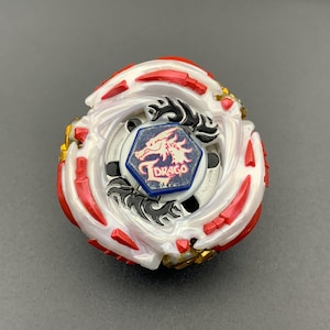May include: A white and red spinning top toy featuring a central hexagonal emblem with a blue background, a red dragon illustration, and the text Drago. The circular toy has metallic gold accents, black decorative patterns, and a layered plastic design.