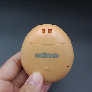May include: A tan, egg-shaped handheld electronic game. The back of the device has a silver label with the number "2147510" printed on it. The device is held in a person's hand.