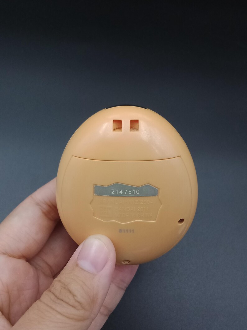 May include: A tan, egg-shaped handheld electronic game. The back of the device has a silver label with the number "2147510" printed on it. The device is held in a person's hand.