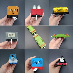 以下が含まれることがあります： カラフルなおもちゃの電車車両と機関車のコレクション。オレンジ色の貨車、赤いミルクタンカー、"SODOR FUEL"の文字が入った黄色の燃料タンカー、灰色の貨車、2つの緑色のフラットベッド車、青いトーマス・ザ・タンク・エンジン、青い車両が含まれています。