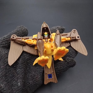 May include: A yellow and brown bird-like action figure with articulated wings and legs. The toy has a detailed design with a bird head and a small blue square on the body. The figure is held in a black glove.