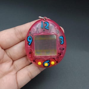 May include: A pink, retro handheld digital pet toy with a small screen and blue numbers. The toy has a keychain loop and features the numbers 12, 3, 6, and 9. Arrows and yellow dots are also visible on the device.