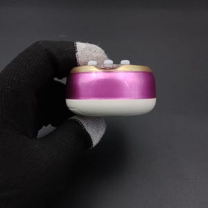 Puede incluir: Un peque&ntilde;o dispositivo port&aacute;til de forma rectangular redondeada. Tiene un cuerpo rosa met&aacute;lico, un borde dorado y una base blanca. Tres peque&ntilde;os botones blancos son visibles en la parte superior. El dispositivo es sostenido por una mano enguantada sobre un fondo oscuro.