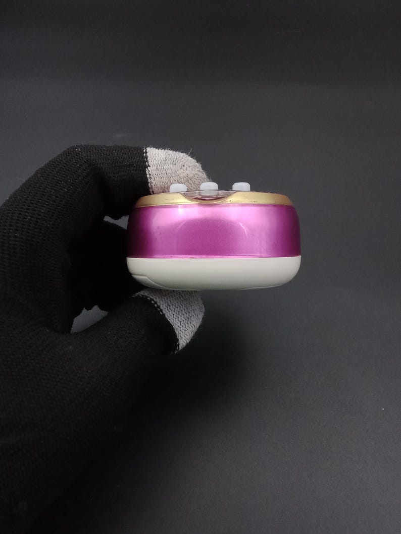 Puede incluir: Un peque&ntilde;o dispositivo port&aacute;til de forma rectangular redondeada. Tiene un cuerpo rosa met&aacute;lico, un borde dorado y una base blanca. Tres peque&ntilde;os botones blancos son visibles en la parte superior. El dispositivo es sostenido por una mano enguantada sobre un fondo oscuro.