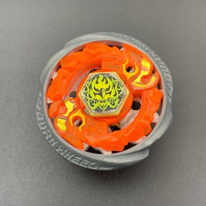 Op de afbeelding: Een cirkelvormig tol-speeltje met een metallic grijze buitenring waarop de tekst BURN WHEEL is gegraveerd. Het midden bestaat uit een doorschijnend oranje kunststof vlamvormig onderdeel rondom een zeshoekige bout met een feniksafbeelding.