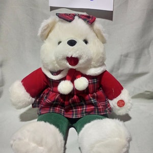 May include: A white plush teddy bear wearing a red and green tartan dress with a white bow. The bear has a red mouth and black eyes. The bear is sitting on a white surface. The bear has a button on its chest that says "Press for Music".