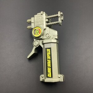 May include: A silver-colored plastic Beyblade Spring Shooter toy with a textured handle grip. The device features a yellow and red circular indicator with the letter R, exposed metal springs, and a black label with yellow text reading BEYBLADE SPRING SHOOTER.