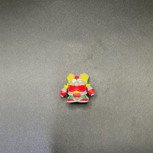 May include: A small, colorful toy robot figurine. The robot has a red body, yellow ears, green and yellow arms, and red hands. The robot is standing on a gray surface.