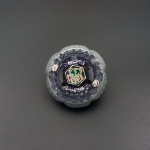 Puede incluir: Un juguete de peonza circular con un anillo exterior de metal plateado con borde festoneado. El centro muestra un emblema circular negro con un gráfico estilizado de cara de tigre en verde y naranja y detalles en morado.