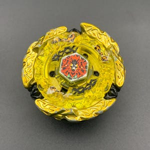 Könnte beinhalten: Ein kreisförmiges gelb-goldenes Kreiselspielzeug mit einem zentralen sechseckigen Emblem mit rotem und schwarzem Grafikdesign. Das Spielzeug hat eine durchscheinende gelbe Kunststoffschicht mit schwarzen Akzenten und einen metallisch goldenen Außenring.