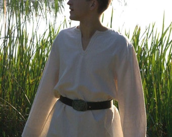 May include: A man wearing a white long-sleeved shirt with a brown belt and black trousers. He is standing in a field of tall green grass.