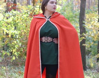 May include: A woman wearing a red hooded cape with white trim, a green shirt, and brown boots. She is standing in a forest with fall foliage.