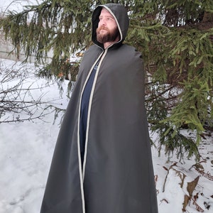 Puede incluir: Una capa con capucha de color verde oscuro con ribete blanco. La capa la lleva puesta una persona de pie en un paisaje nevado.