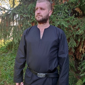 May include: A black long-sleeved shirt with a V-neck and a leather belt with a silver buckle. The shirt is being worn by a person with a beard standing in front of a green tree.