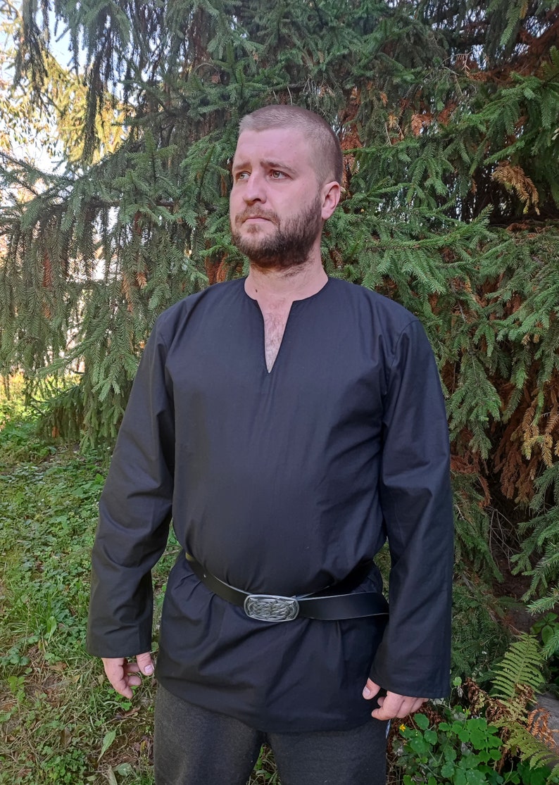 May include: A black long-sleeved shirt with a V-neck and a leather belt with a silver buckle. The shirt is being worn by a person with a beard standing in front of a green tree.
