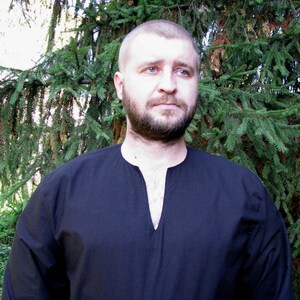 May include: A man wearing a black long-sleeved shirt with a V-neck. He has a beard and short brown hair. He is standing in front of a green tree.