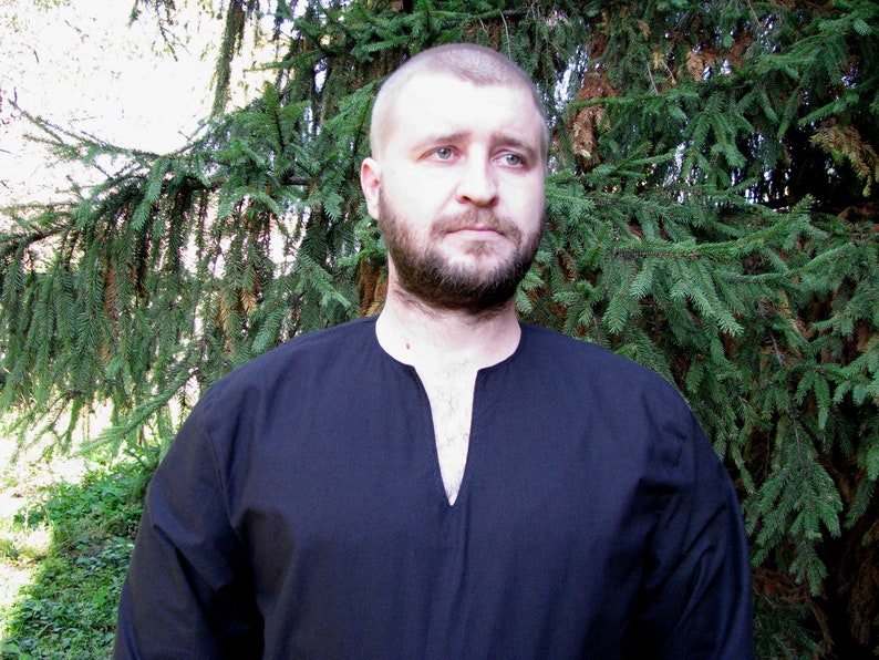 May include: A man wearing a black long-sleeved shirt with a V-neck. He has a beard and short brown hair. He is standing in front of a green tree.
