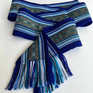 Peut inclure: Une ceinture tissée bleue avec un motif géométrique et des franges. La ceinture est pliée et les franges sont visibles.