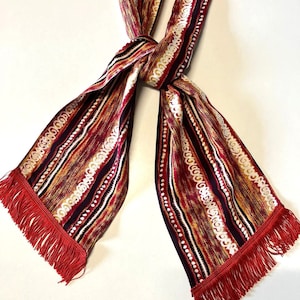 May include: A red, white, and gold striped scarf with a fringe on each end. The scarf has a geometric pattern.
