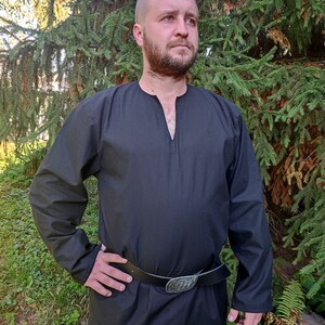 May include: A black long-sleeved shirt with a V-neck and a black leather belt with a silver buckle. The shirt is being worn by a person standing in front of a green background.