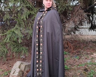 May include: A long, dark brown hooded cloak with a decorative trim of crocheted flowers in shades of brown, orange, and white. The cloak is worn by a person with blonde hair.