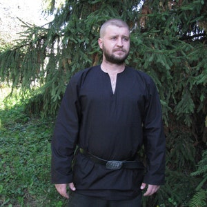 May include: A man wearing a black long-sleeved shirt with a v-neck and a black leather belt with a silver buckle. He is standing in front of a green pine tree.