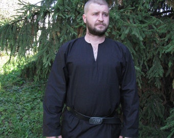 Op de afbeelding: Een man draagt een zwart lang mouw shirt met een v-hals en een zwarte leren riem met een zilveren gesp. Hij staat voor een groene dennenboom.