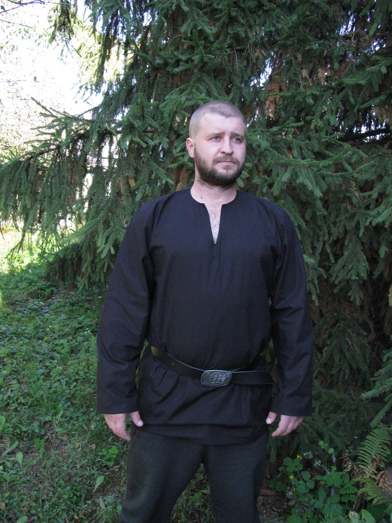 May include: A man wearing a black long-sleeved shirt with a v-neck and a black leather belt with a silver buckle. He is standing in front of a green pine tree.
