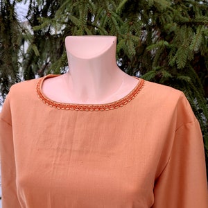 K&ouml;nnte beinhalten: Eine orangefarbene Tunika mit langen &Auml;rmeln und einem dekorativen Besatz am Ausschnitt. Der Besatz ist aus einem gewebten Stoff mit einem geometrischen Muster.
