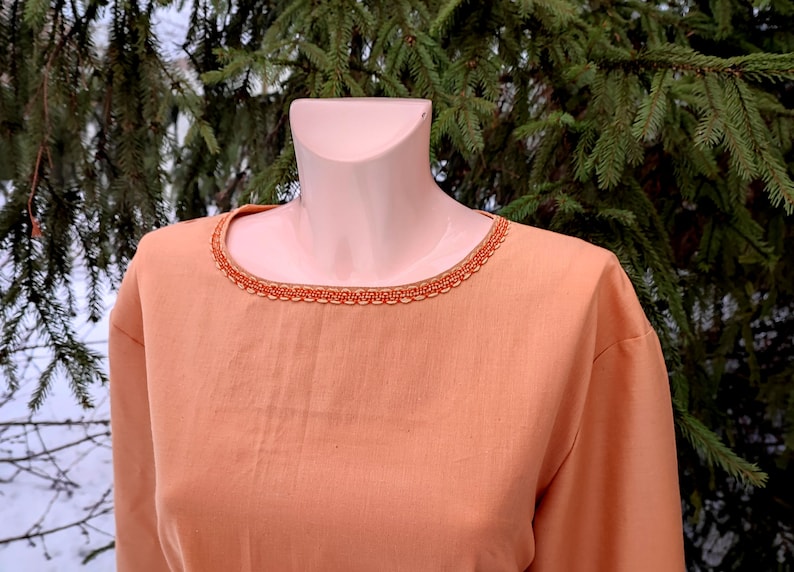 K&ouml;nnte beinhalten: Eine orangefarbene Tunika mit langen &Auml;rmeln und einem dekorativen Besatz am Ausschnitt. Der Besatz ist aus einem gewebten Stoff mit einem geometrischen Muster.