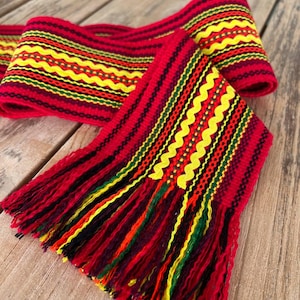 Puede incluir: Un cinturón rojo tejido con patrones de zigzag y rayas amarillas, verdes y negras. El cinturón tiene largas y coloridas borlas en rojo, amarillo, verde, naranja y negro. El cinturón se muestra sobre una superficie de madera.