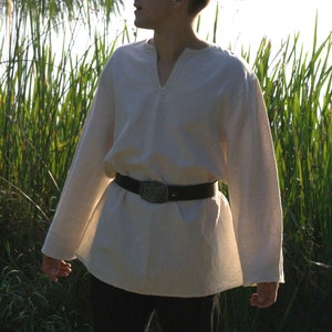 May include: A man wearing a white linen shirt with a brown leather belt and a silver buckle. He is standing in a field of tall grass.