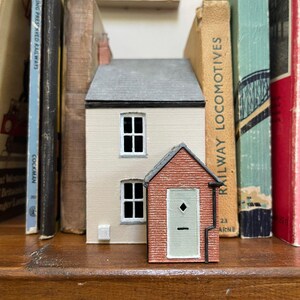 May include: A miniature model of a two-story house with cream-colored walls and a gray roof, positioned next to a smaller red brick outbuilding featuring a light green door with a diamond-shaped window, set between several vintage hardcover books.