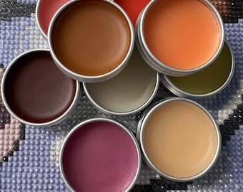 Könnte beinhalten: Eine Sammlung von neun runden silbernen Metalldosen, gefüllt mit verschiedenen Farbtönen von festem Cremepigment. Die Farben umfassen Dunkelbraun, leuchtendes Rot, Orange, helles Beige, Olivgrün, gedämpftes Salbei, Dunkelbeere und sanfte Rosatöne.