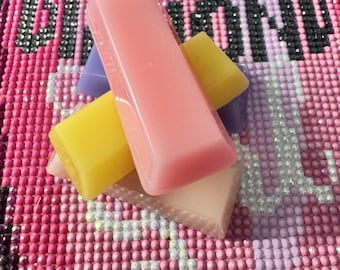 Könnte beinhalten: Ein Stapel rechteckiger Wachsstücke in den Farben Rosa, Gelb und Violett. Die Wachsstücke haben eine glatte, feste Oberfläche und sind in einer Gruppe angeordnet, die auf einem Hintergrund mit einem rosa Diamant-Kunstmuster und dem Wort DIAMOND liegt.