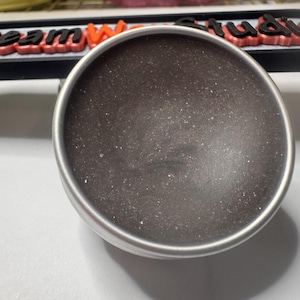 May include: A circular, metallic container with a dark, speckled interior. The container has a silver rim and rests on a white surface. In the background, a sign reads "Dream World Studio" in red and black.