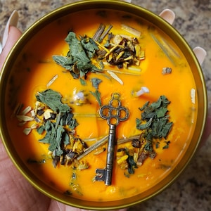 May include: A gold candle jar with an orange wax base and a key charm. The wax is filled with dried herbs and flowers.
