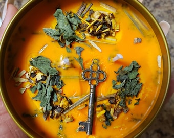 May include: A gold candle jar with an orange wax base and a key charm. The wax is filled with dried herbs and flowers.