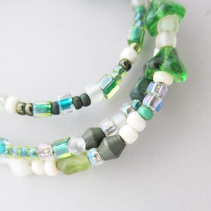 May include: A multi-strand beaded bracelet featuring a variety of glass and matte beads in shades of forest green, lime, white, and iridescent teal. The design incorporates translucent mushroom-shaped beads, small round spheres, and faceted cube beads arranged in a repeating pattern along the flexible wire structure.