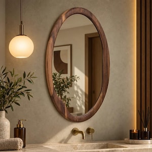 May include: An oval wall mirror with a dark brown wooden frame featuring an irregular, organic edge. The mirror reflects a room with a doorway and a framed picture. It is mounted on a textured wall above a sink with a brass faucet and handle.