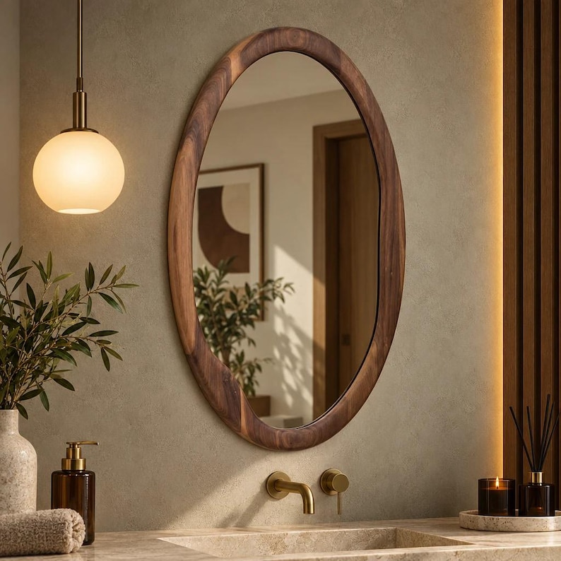 May include: An oval wall mirror with a dark brown wooden frame featuring an irregular, organic edge. The mirror reflects a room with a doorway and a framed picture. It is mounted on a textured wall above a sink with a brass faucet and handle.