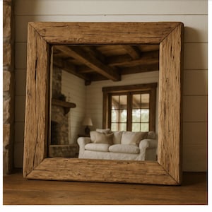 Può includere: Specchio quadrato con cornice in legno rustico. La cornice è realizzata in legno grezzo, marrone chiaro. Lo specchio riflette un soggiorno con un divano bianco, un camino e una finestra. Lo specchio è appoggiato su una superficie di legno.