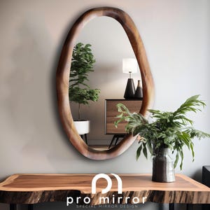 May include: Oval-shaped wooden mirror with a natural, organic edge. The mirror reflects a room with a wooden console table, a lamp, and greenery. The table has a live edge design. The text "pro mirror special mirror design" is at the bottom.