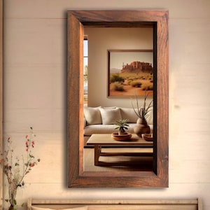 Pode incluir: Um espelho retangular com moldura de madeira marrom. O espelho reflete uma sala de estar com um sofá, uma mesa de centro e uma imagem emoldurada na parede.