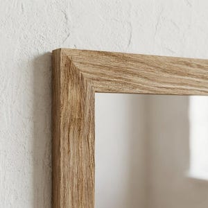 May include: Close-up of a rectangular mirror with a light brown, textured wooden frame. The frame has a natural wood grain appearance and is set against a white textured wall. The mirror reflects a soft, neutral background.