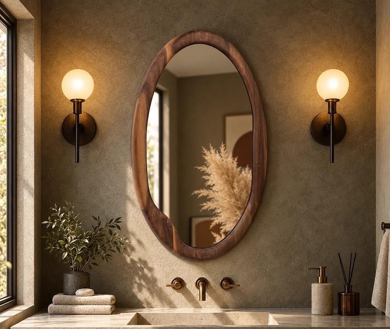 May include: An oval-shaped mirror with a dark brown wooden frame mounted on a textured wall. The mirror is flanked by two spherical wall sconces with warm glowing bulbs. Below the mirror, a faucet and sink are visible with a plant and towels.