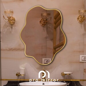 May include: A gold-framed, wavy-shaped mirror hangs on a white tiled wall in a bathroom. The mirror reflects a gold towel rack and other bathroom fixtures.