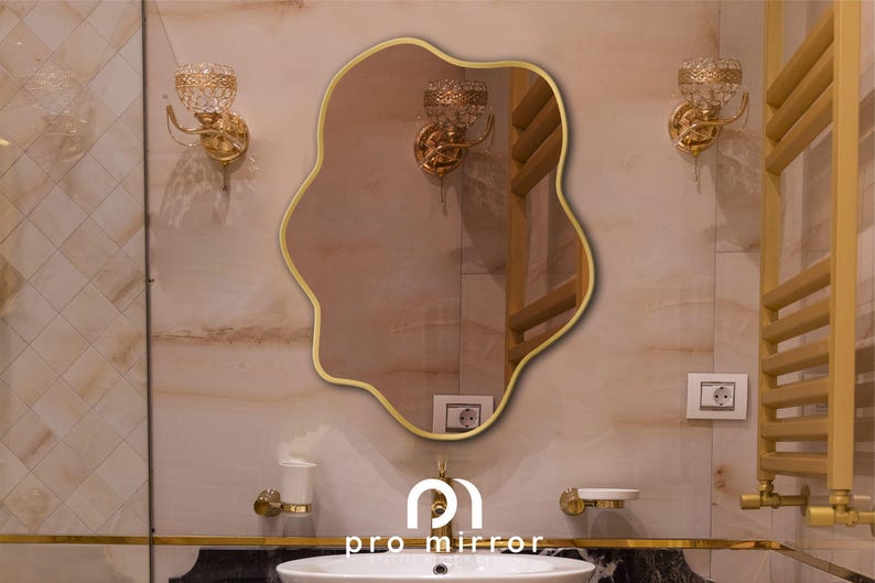 May include: A gold-framed, wavy-shaped mirror hangs on a white tiled wall in a bathroom. The mirror reflects a gold towel rack and other bathroom fixtures.