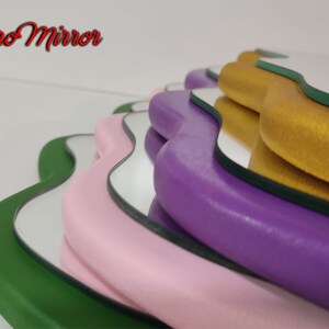 May include: A set of five wavy-edged mirrors with different colored frames. The frames are green, pink, purple, gold, and green. The mirrors are stacked on top of each other.